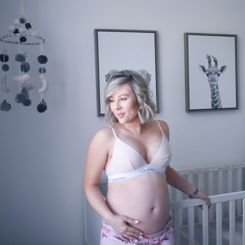 My Cancer Chic Blogger Anna Crollman shares her second trimester experiences - in photo woman standing near a crib and wearing a bra and shorts showing her belly 